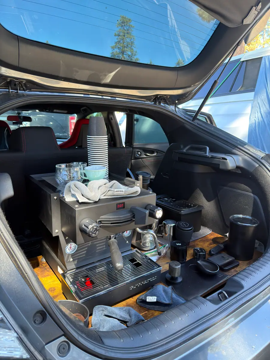 The espresso setup inside the open hatch: a black La Marzocco Linea Mini on a wood counter, a stack of striped cups, a milk pitcher and a tamping station, with the red Type R seats beyond